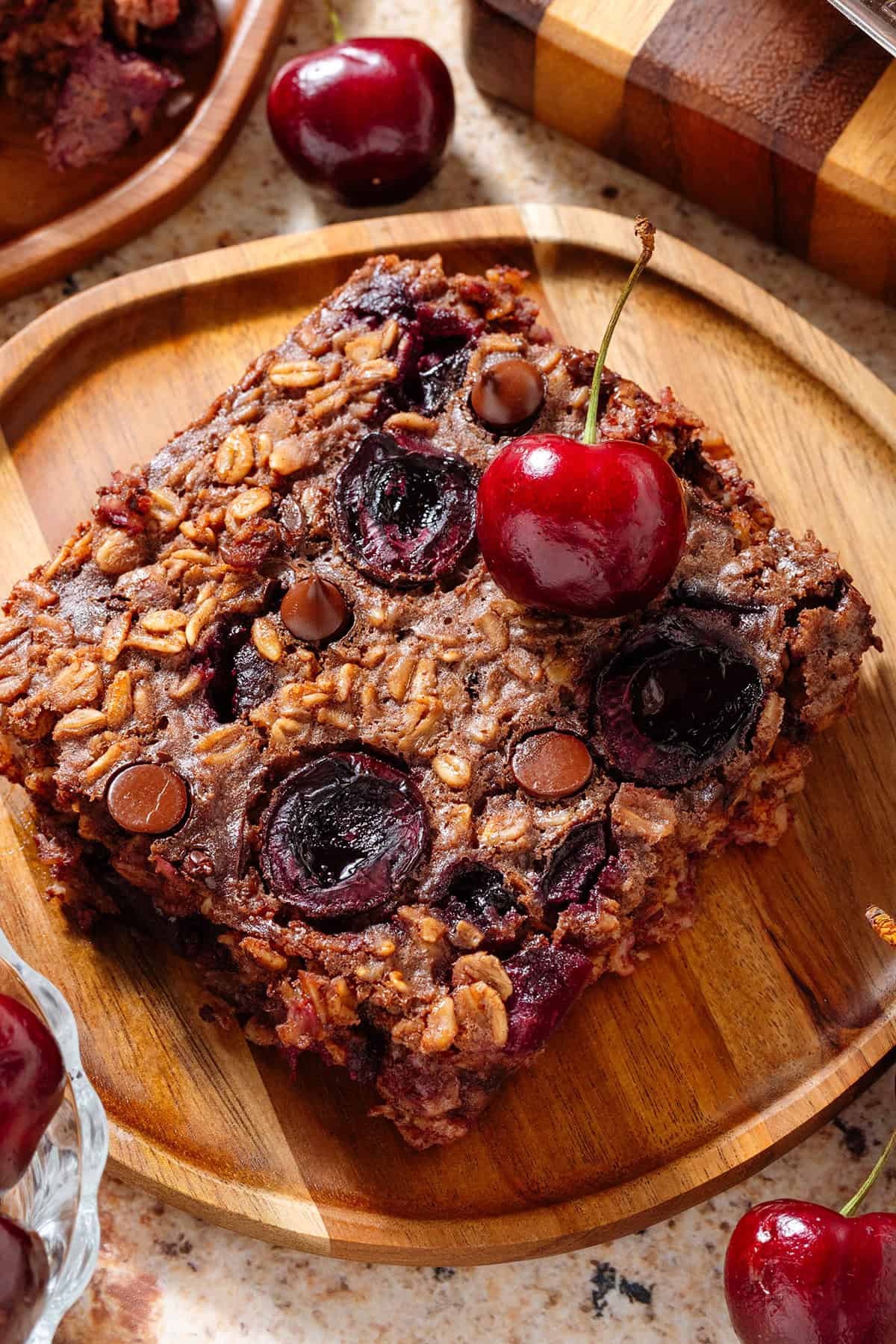 A slice of a chocolate cherry baked oatmeal on a small wooden plate garnished with a fresh cherry.