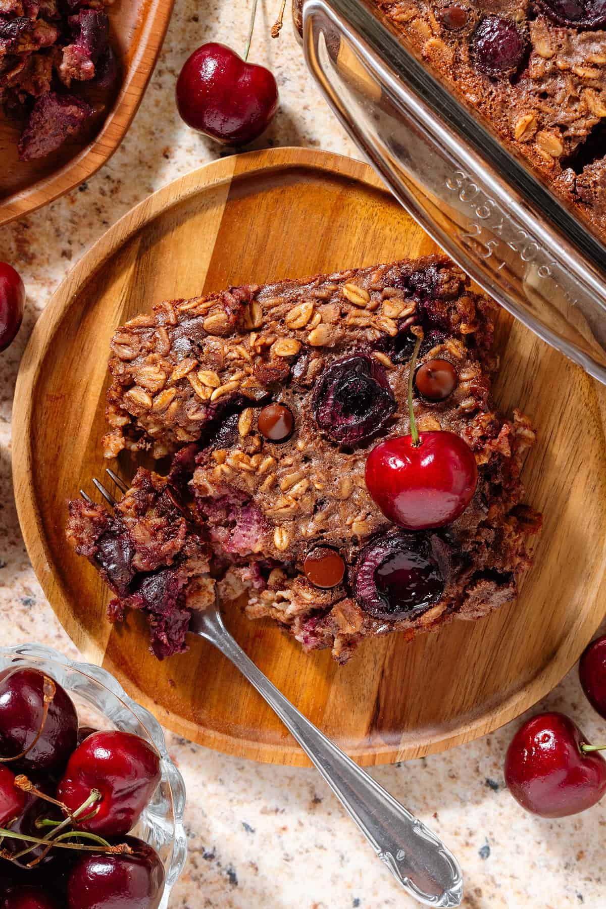 A slice of a chocolate cherry baked oatmeal on a small wooden plate garnished with a fresh cherry and with a fork scooping up a piece of one corner.