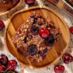 A slice of a chocolate cherry baked oatmeal on a small wooden plate garnished with a fresh cherry and more cherries around.