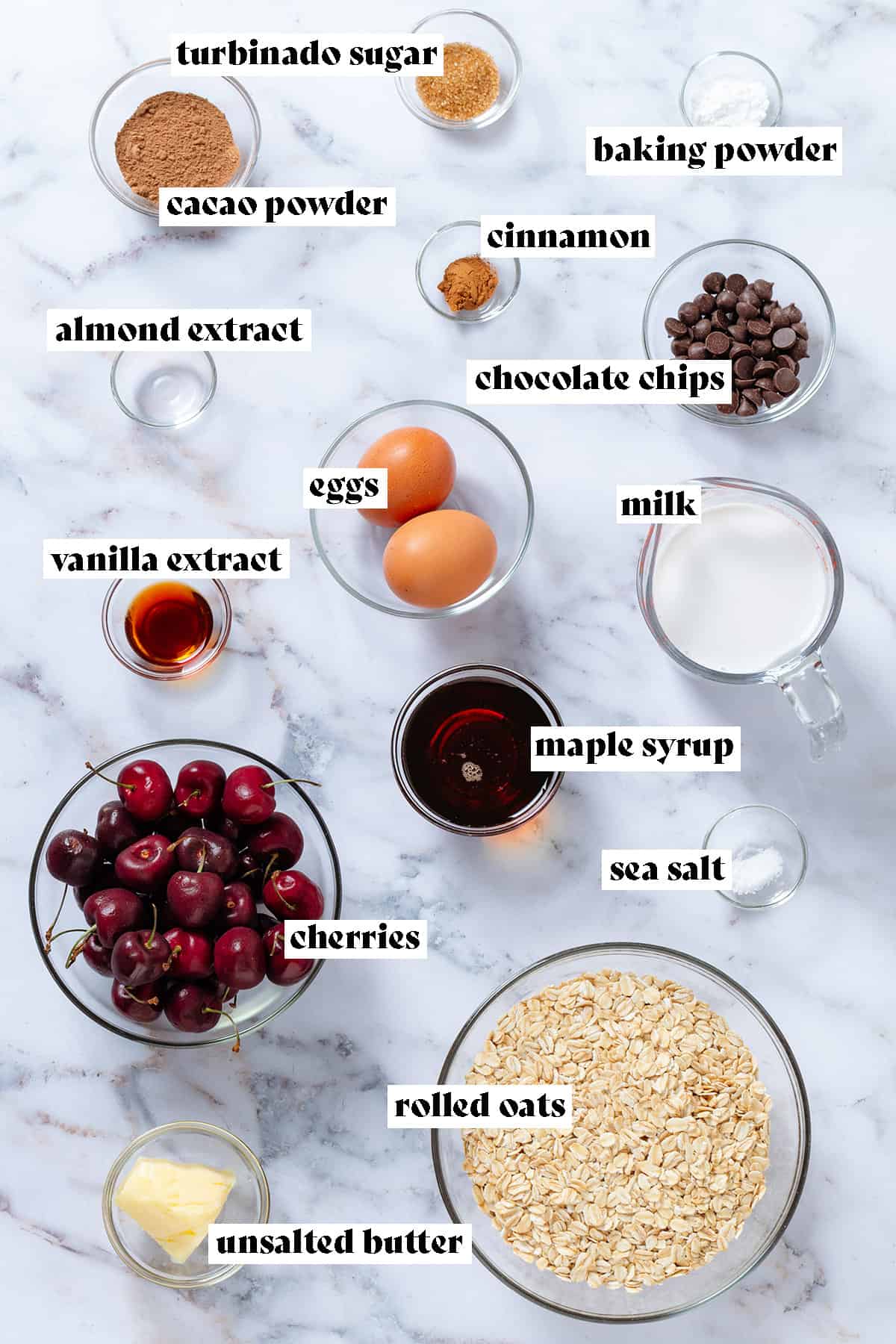 Ingredients in glass bowls like cherries, oats, milk, eggs, chocolate chips, maple syrup, and cacao powder with text overlay.
