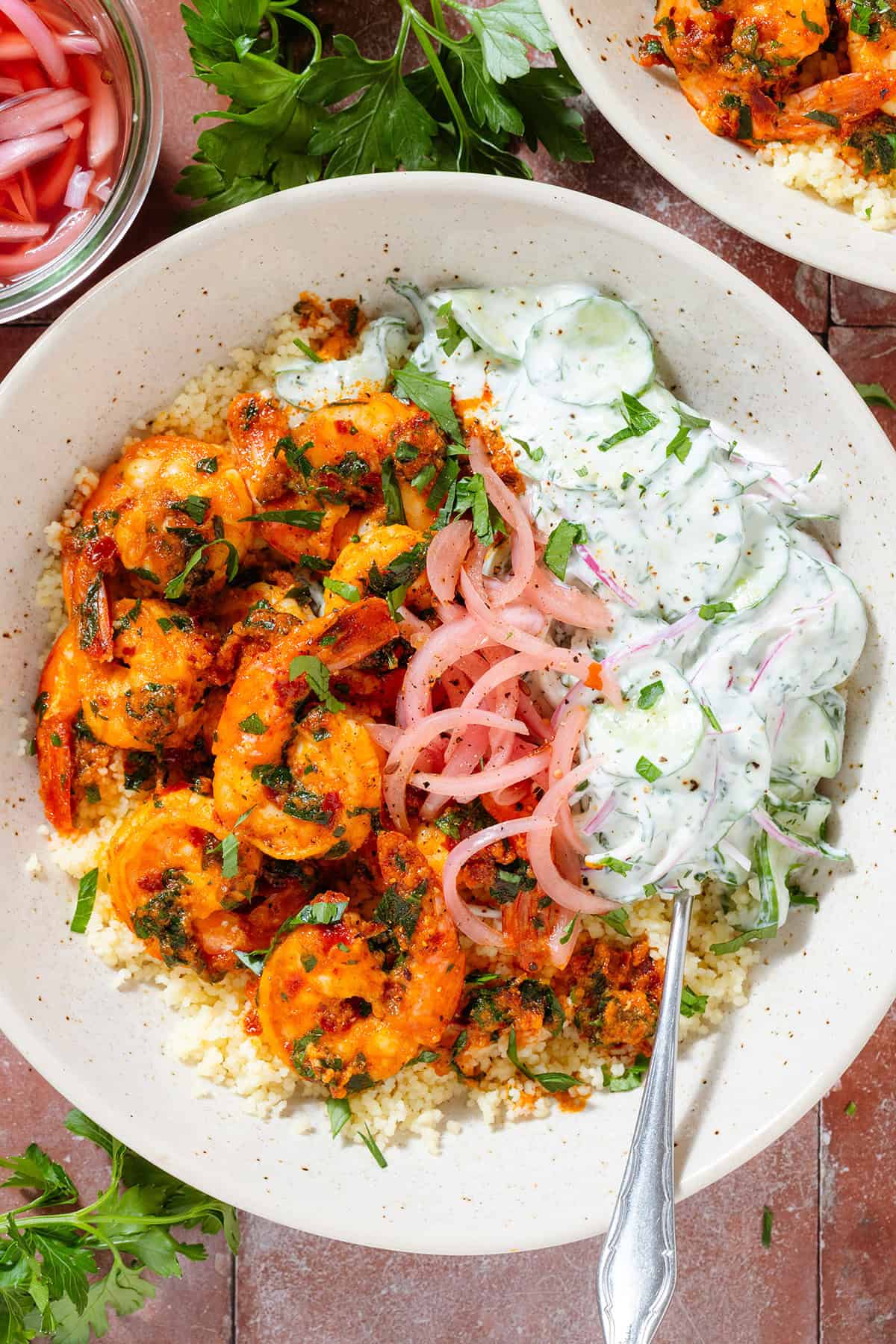 Bright orange harissa shrimp in a beige bowl atop Moroccan couscous, with creamy cucumber salad and pickled red onion.