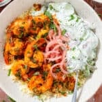 A bowl with harissa shrimp served on Moroccan couscous, with creamy cucumber salad and pickled red onion.