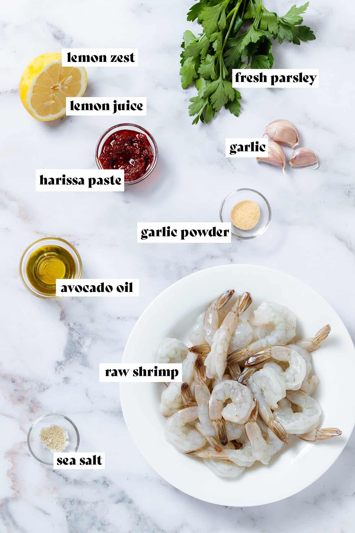 Raw shrimp in a white bowl, harissa, oil, and spices in small glass bowls, fresh parsley, garlic, and lemon all laid out with text overlay.