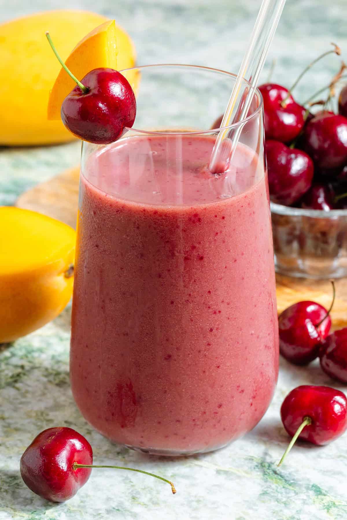 A mango cherry smoothie in a tall glass with a glass straw garnished with a fresh cherry and a slice of mango with more fruit in the background.