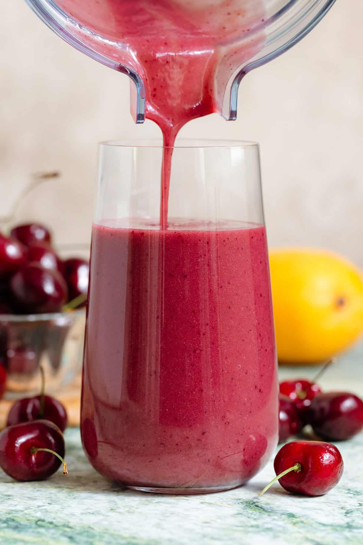 A dark pink smoothie being poured into a tall glass with cherries around it.