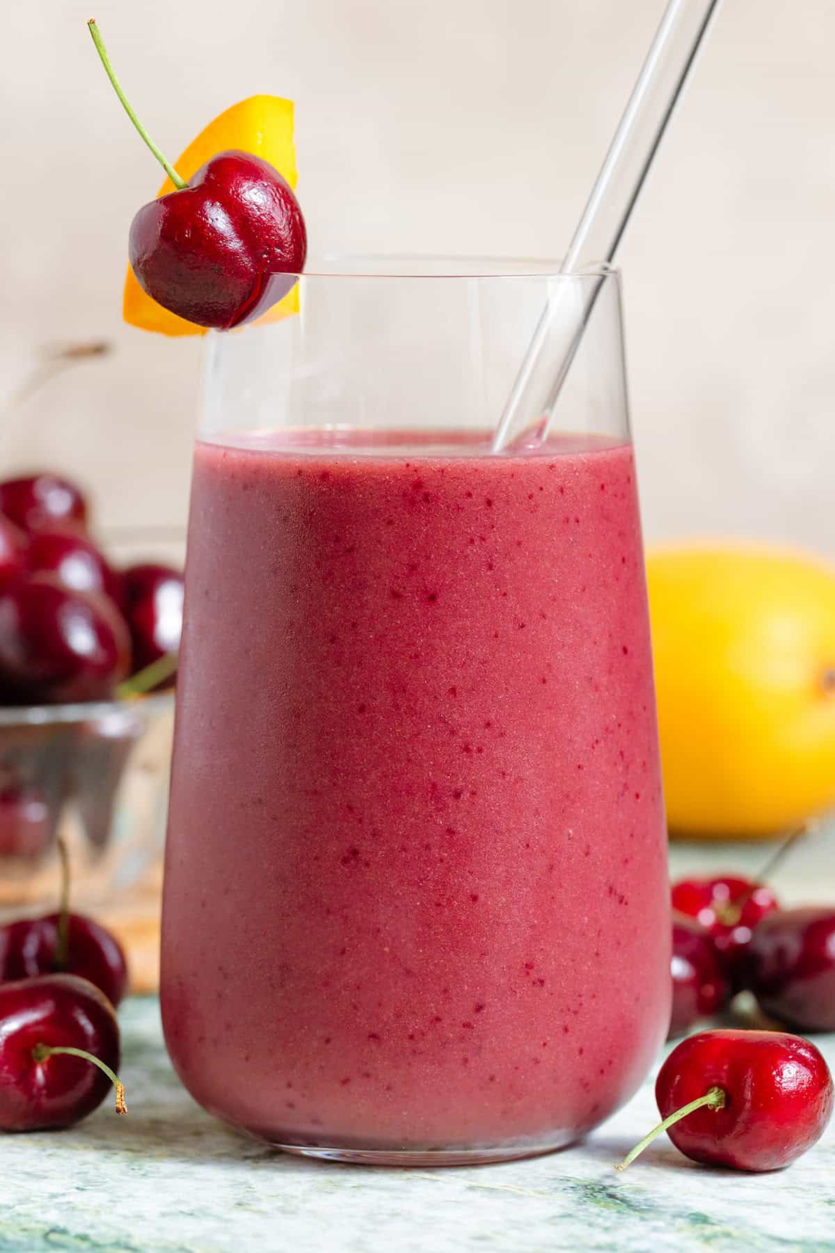 A mango cherry smoothie in a tall glass with a glass straw garnished with a fresh cherry and a slice of mango with more fruit in the background.