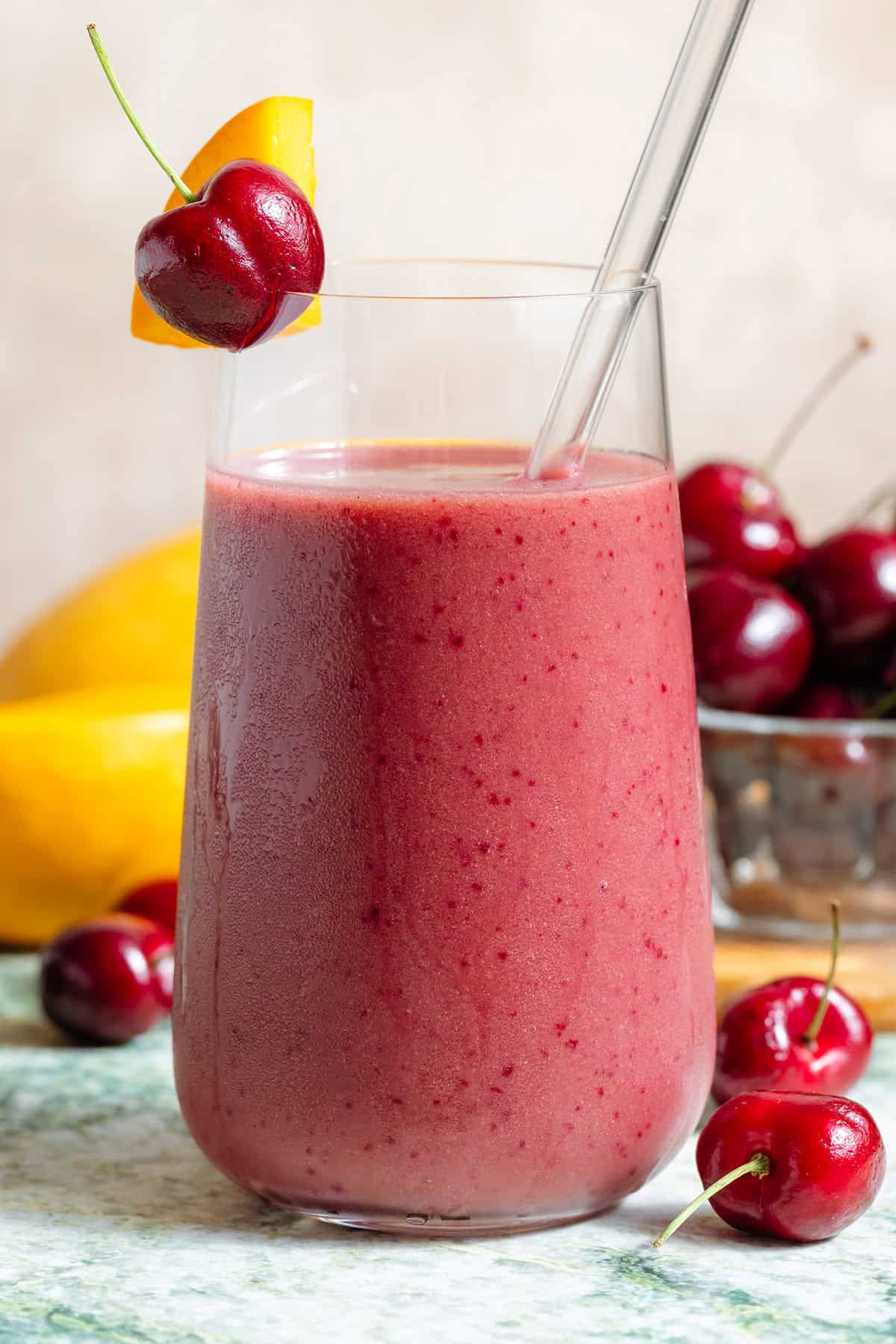 A mango cherry smoothie in a tall glass with a glass straw garnished with a fresh cherry and a slice of mango with more fruit in the background.