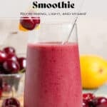 A mango cherry smoothie in a tall glass with a glass straw garnished with a fresh cherry and a slice of mango with more of the fruit in the background.