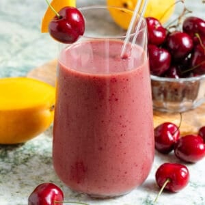 A mango cherry smoothie in a tall glass with a glass straw garnished with a fresh cherry and a slice of mango with more of the fruit in the background.