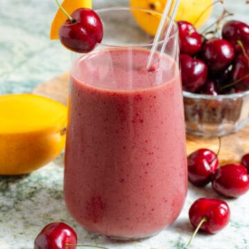 A mango cherry smoothie in a tall glass with a glass straw garnished with a fresh cherry and a slice of mango with more of the fruit in the background.