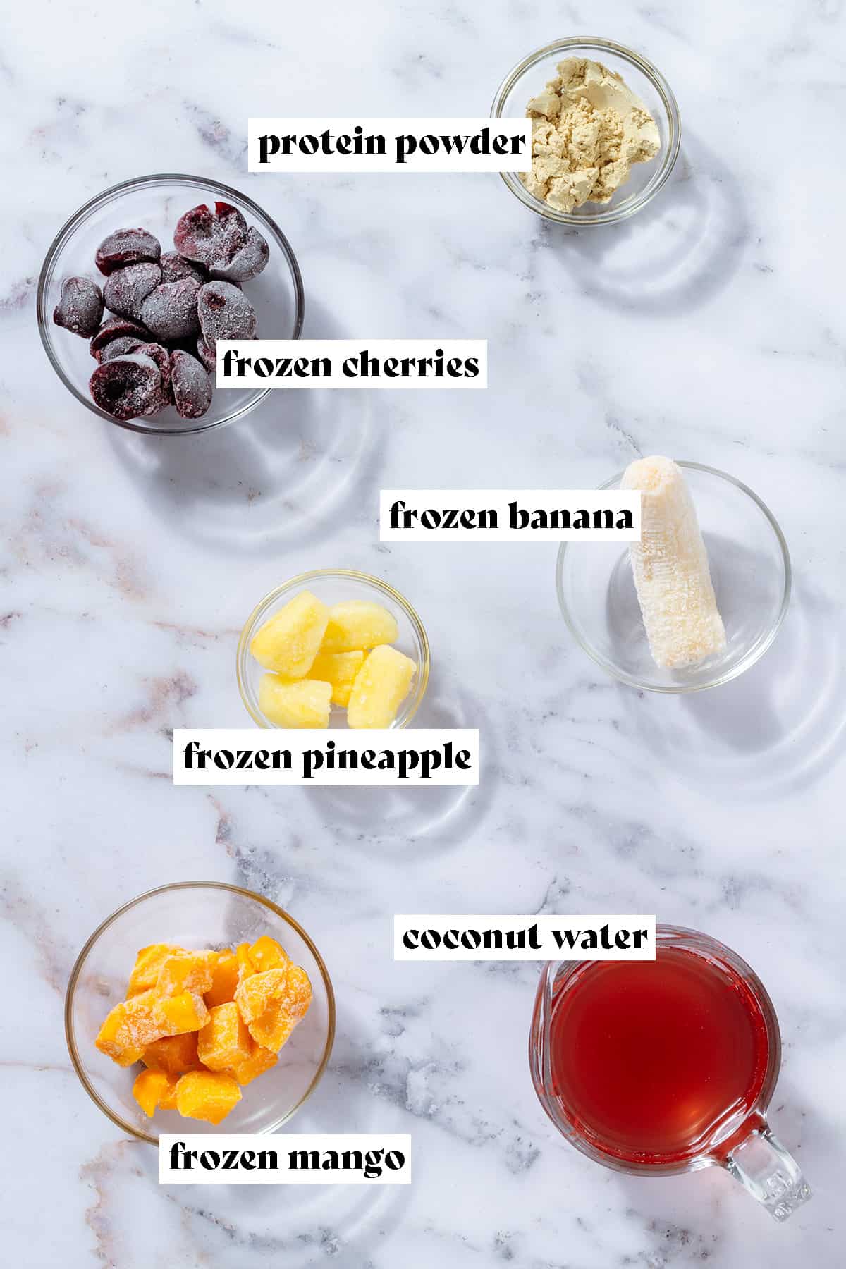 Glass bowls with ingredients like frozen cherries, banana, pineapple, mango, protein powder, and a glass measuring cup with coconut water.