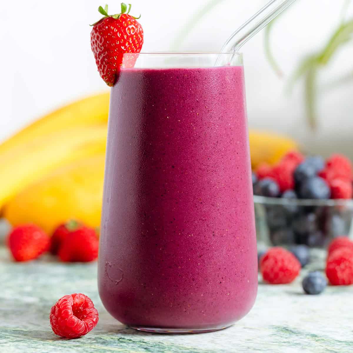 A tall glass with bright purple maqui berry smoothie and a glass straw garnished with a strawberry.