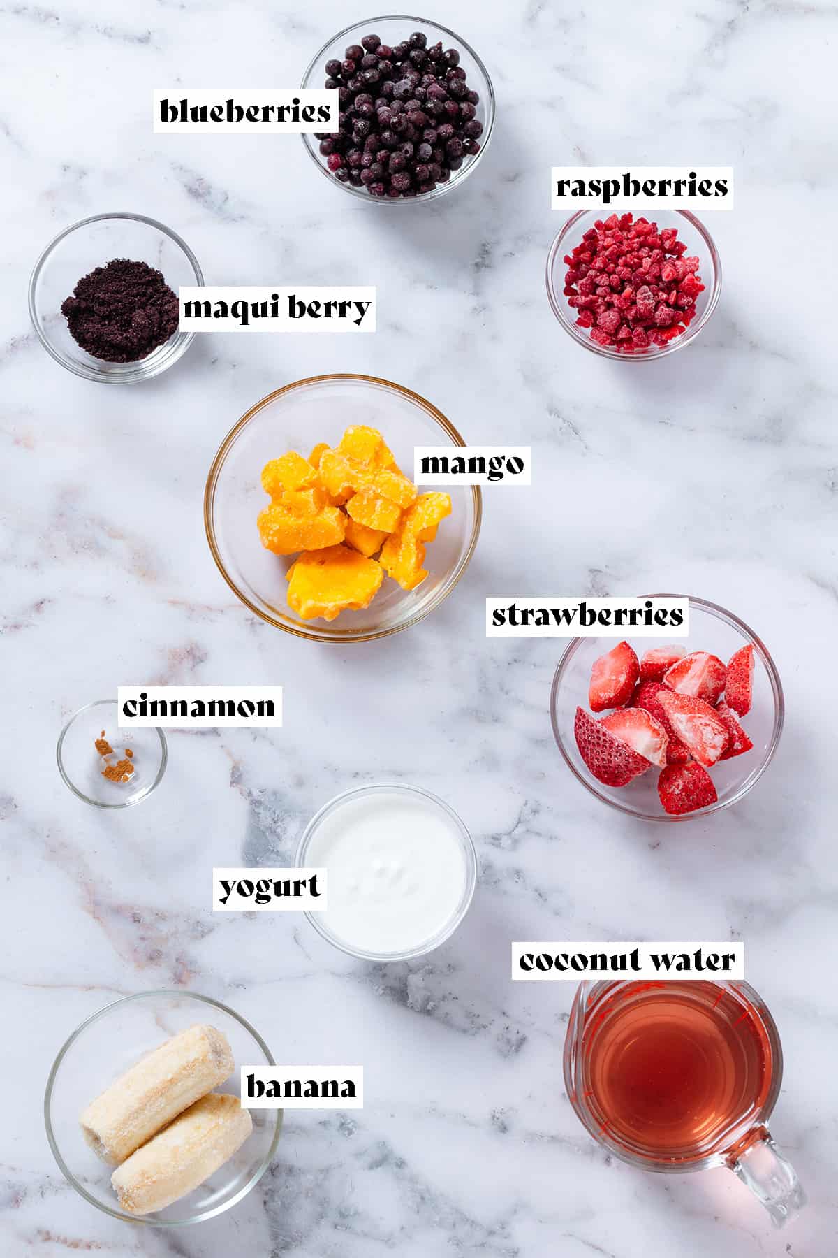 Ingredients in small glass bowls like frozen berries, mango, banana, maqui powder, yogurt, and coconut water.