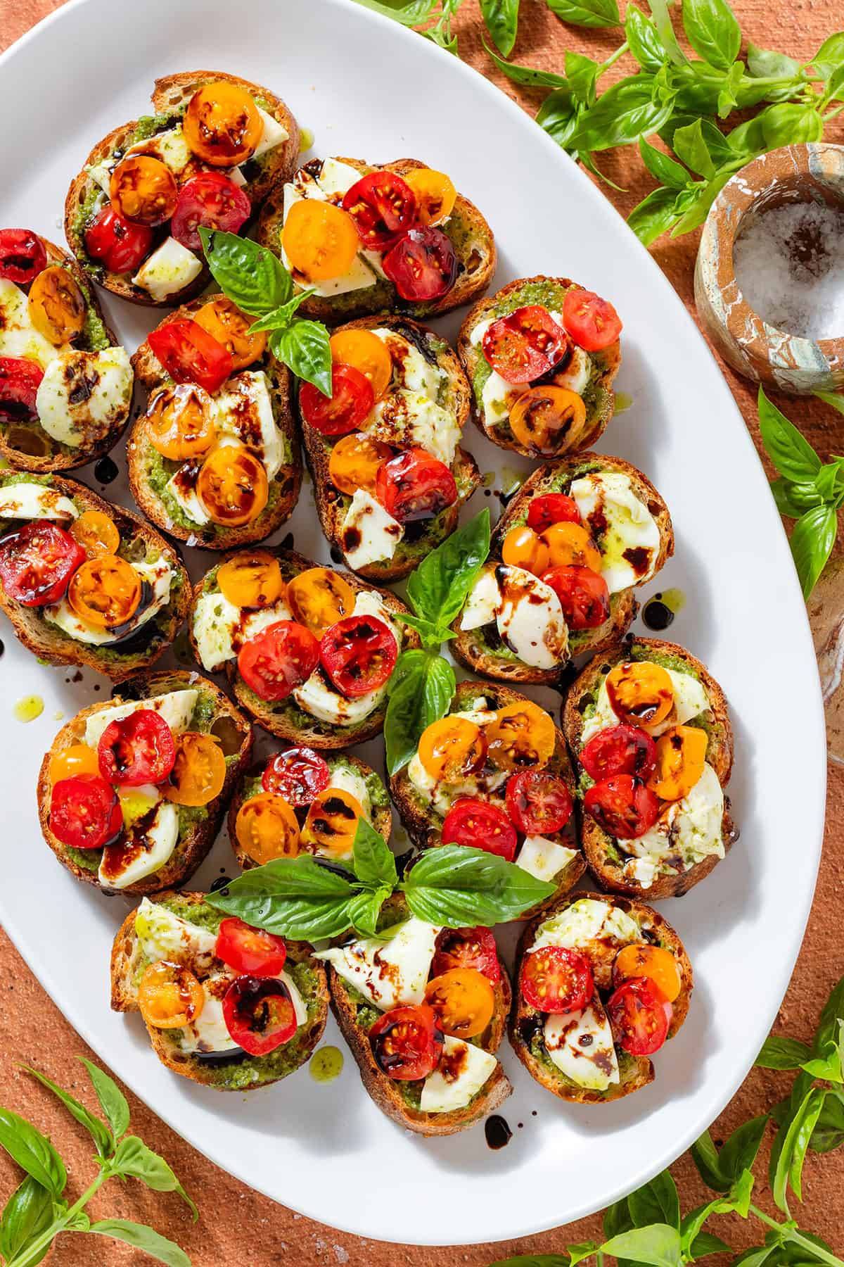 Crostini with pesto, mozzarella, cherry tomatoes drizzled with balsamic glaze and garnished with fresh basil leaves on a large white serving plate.