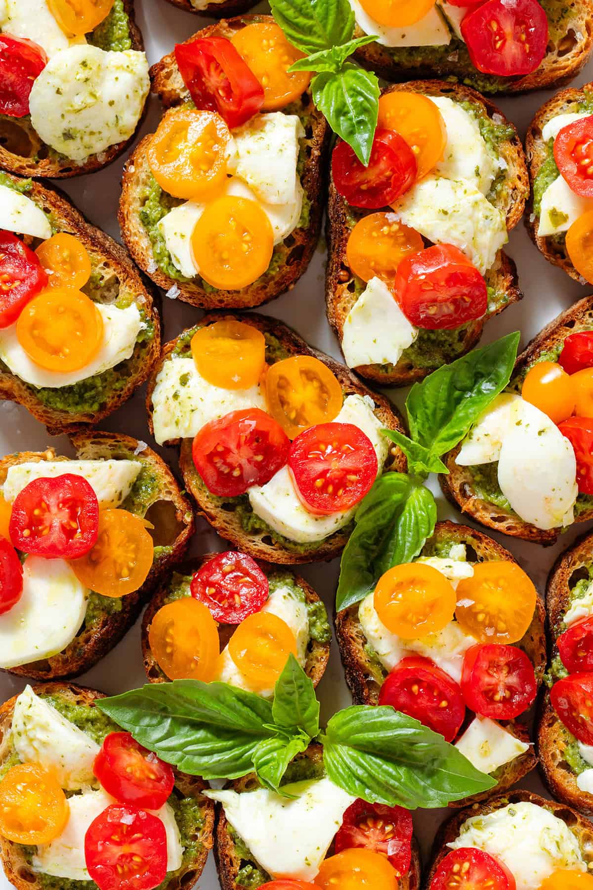 A close up of crostini slices with pesto, mozzarella, cherry tomatoes sprinkled with flaky sea salt.