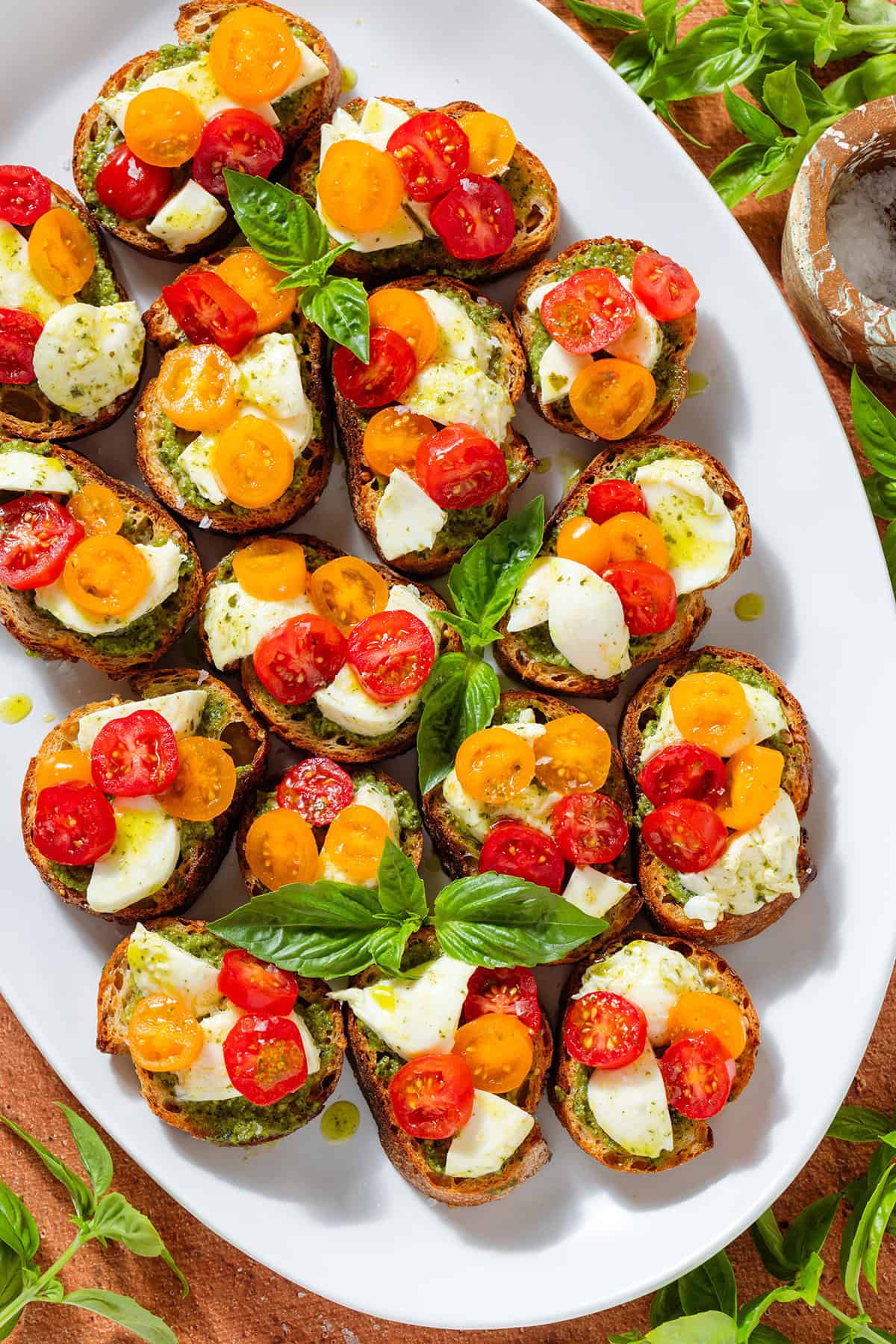 Crostini with pesto, mozzarella, cherry tomatoes garnished with fresh basil leaves on a large white serving plate.