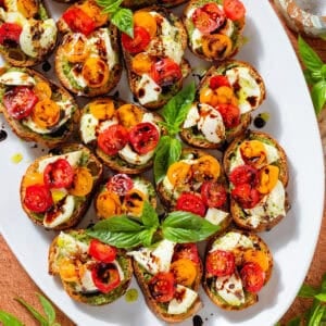 Crostini with pesto, mozzarella, cherry tomatoes drizzled with balsamic glaze and garnished with fresh basil leaves on a large white plate.