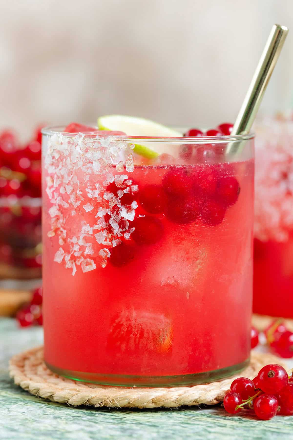 Vibrant red margarita in a short glass with ice garnished with a salt rim on one side, red currants and a lime slice.