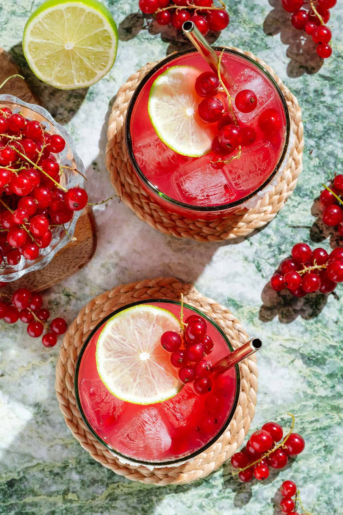Vibrant red margaritas in a short glasses with ice and gold straws garnished with red currants and lime slices.