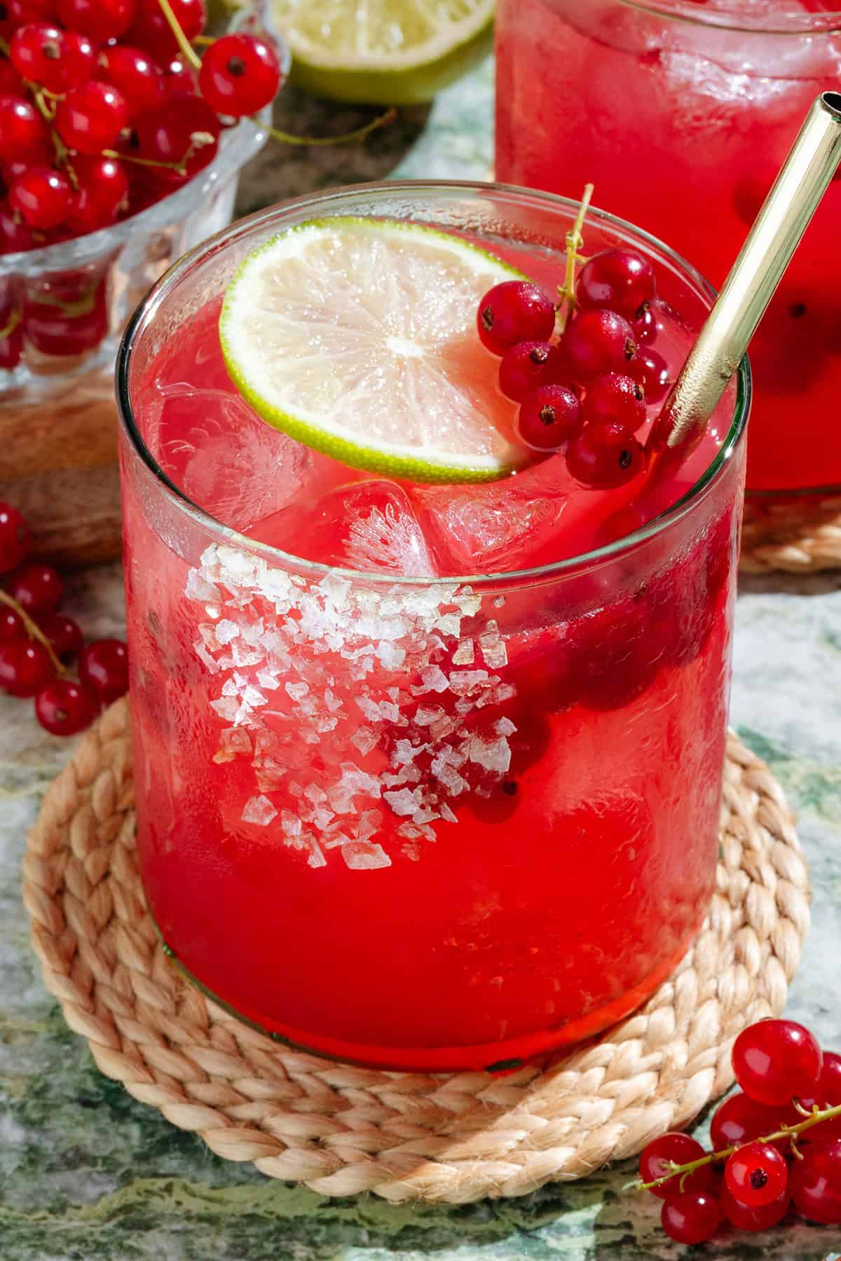 Vibrant red margarita in a short glass with ice and a gold straw garnished with a salt rim, red currants and a lime slice.
