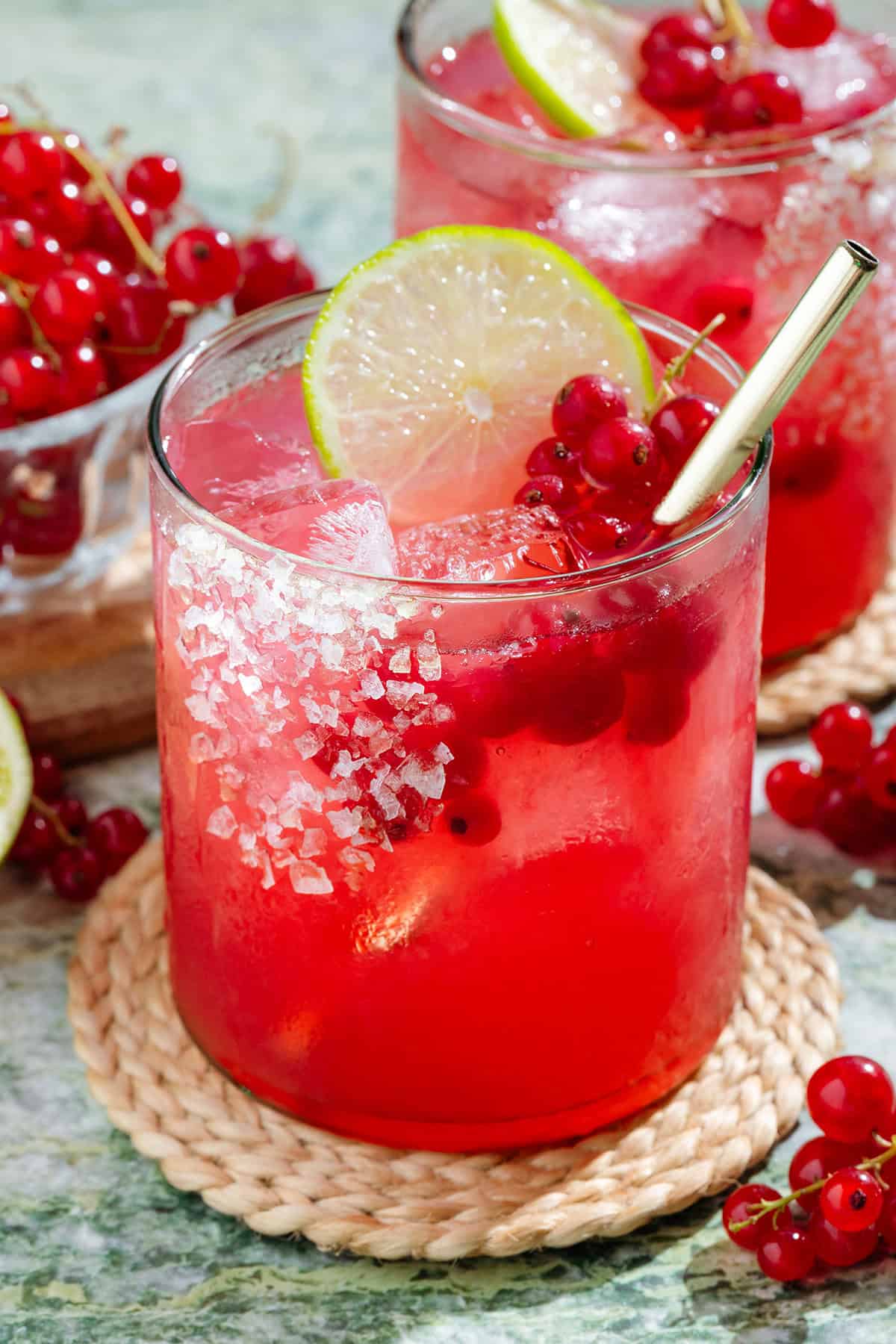 Vibrant red margarita in a short glass with ice and a gold straw garnished with red currants and a lime slice.