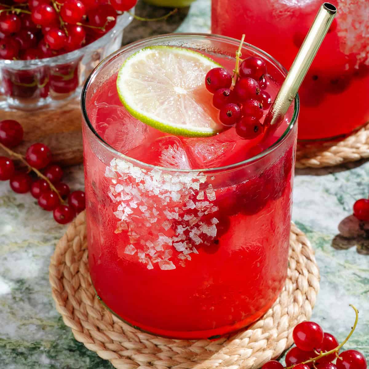 Vibrant red margarita in a short glass with ice garnished with a slice of lime and red currants, with a gold straw.