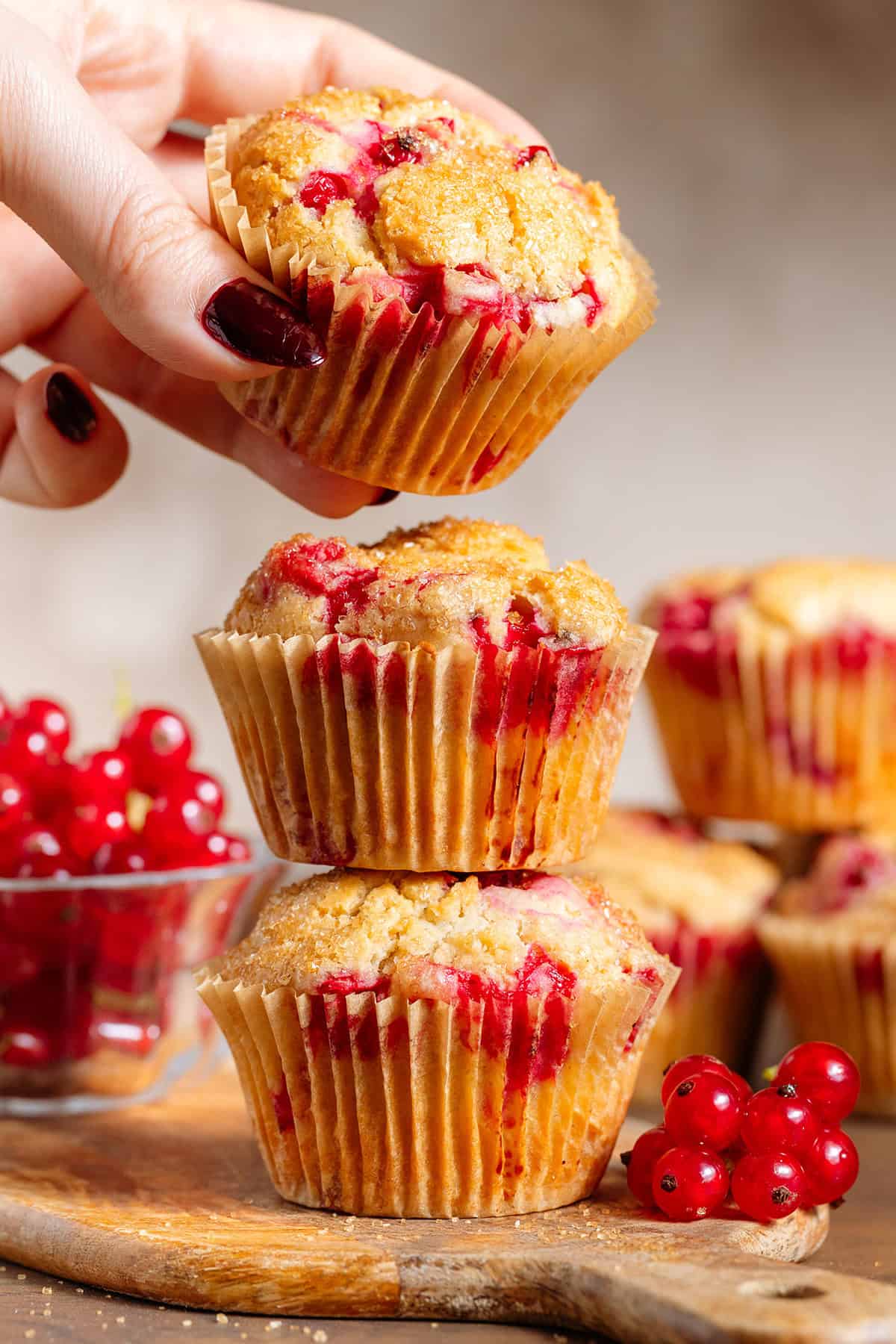 Three red currant muffins stacked on a small wooden cutting board with a hand grabbing the top one.