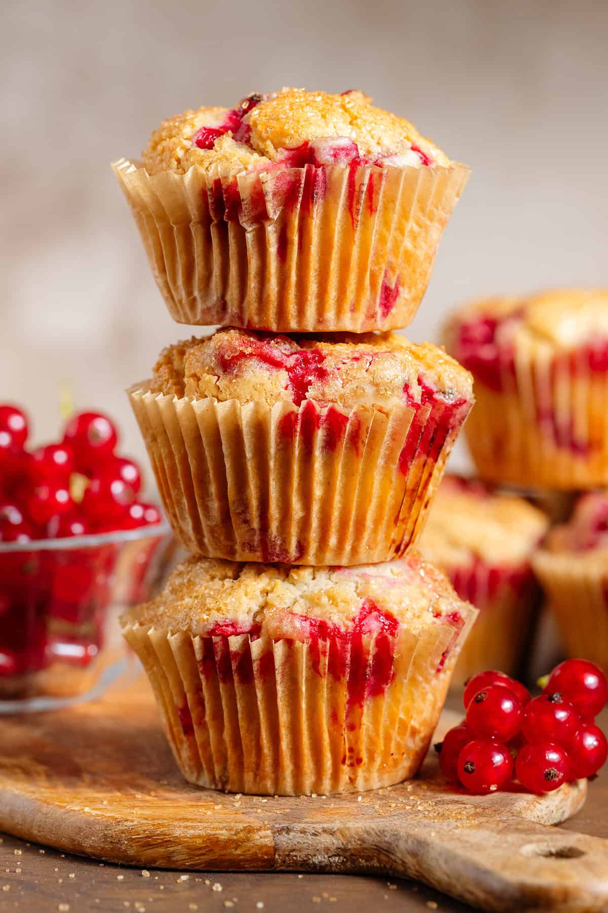 Three red currant muffins stacked on a small wooden cutting board with fresh red currants around and more muffins in the back.