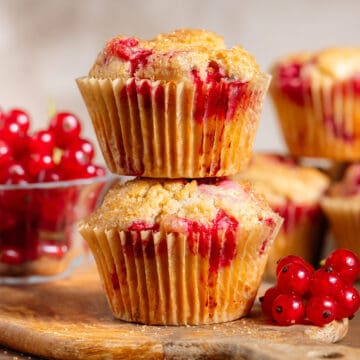 Two red currant muffins stacked on a small wooden cutting board with fresh red currants around and more muffins in the back.