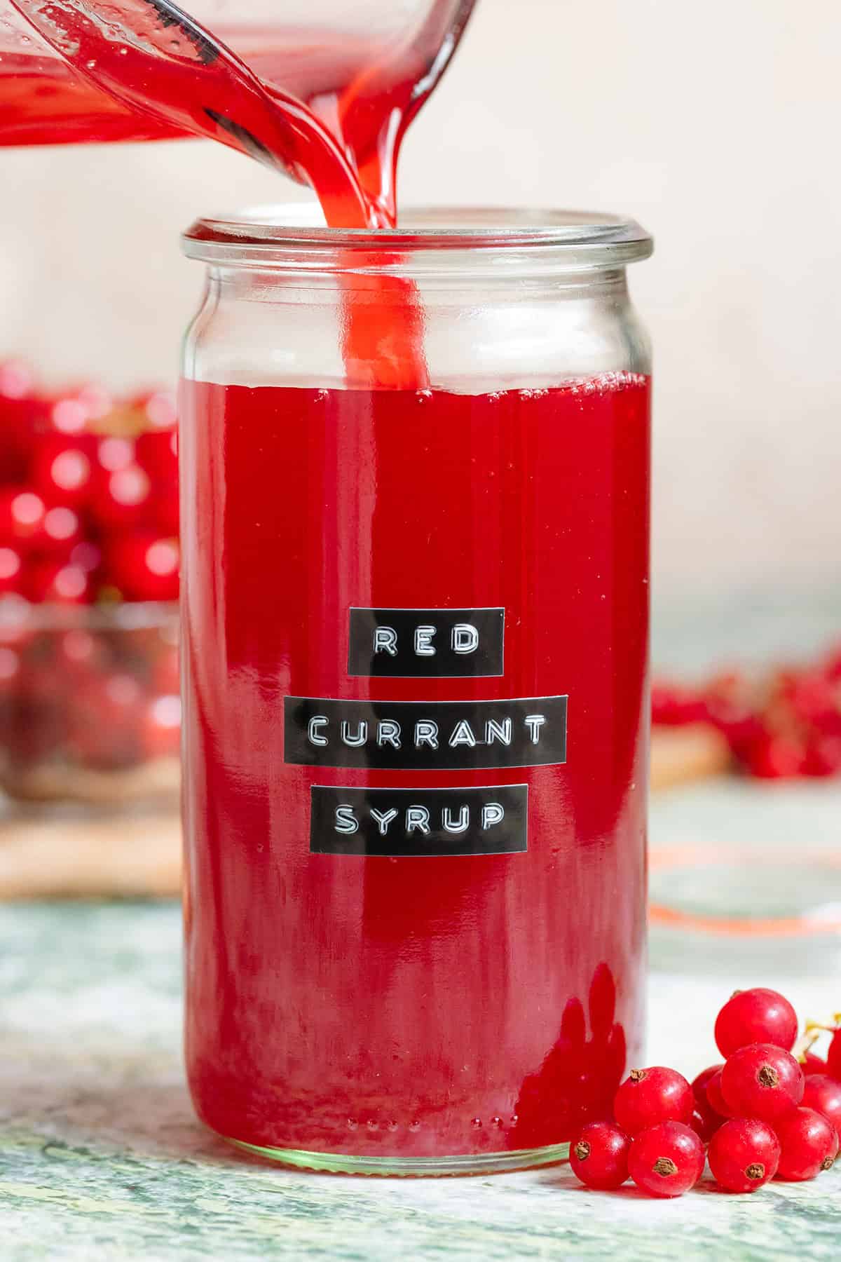 Vibrant red currant syrup being poured into a tall glass jar with an embossed label.