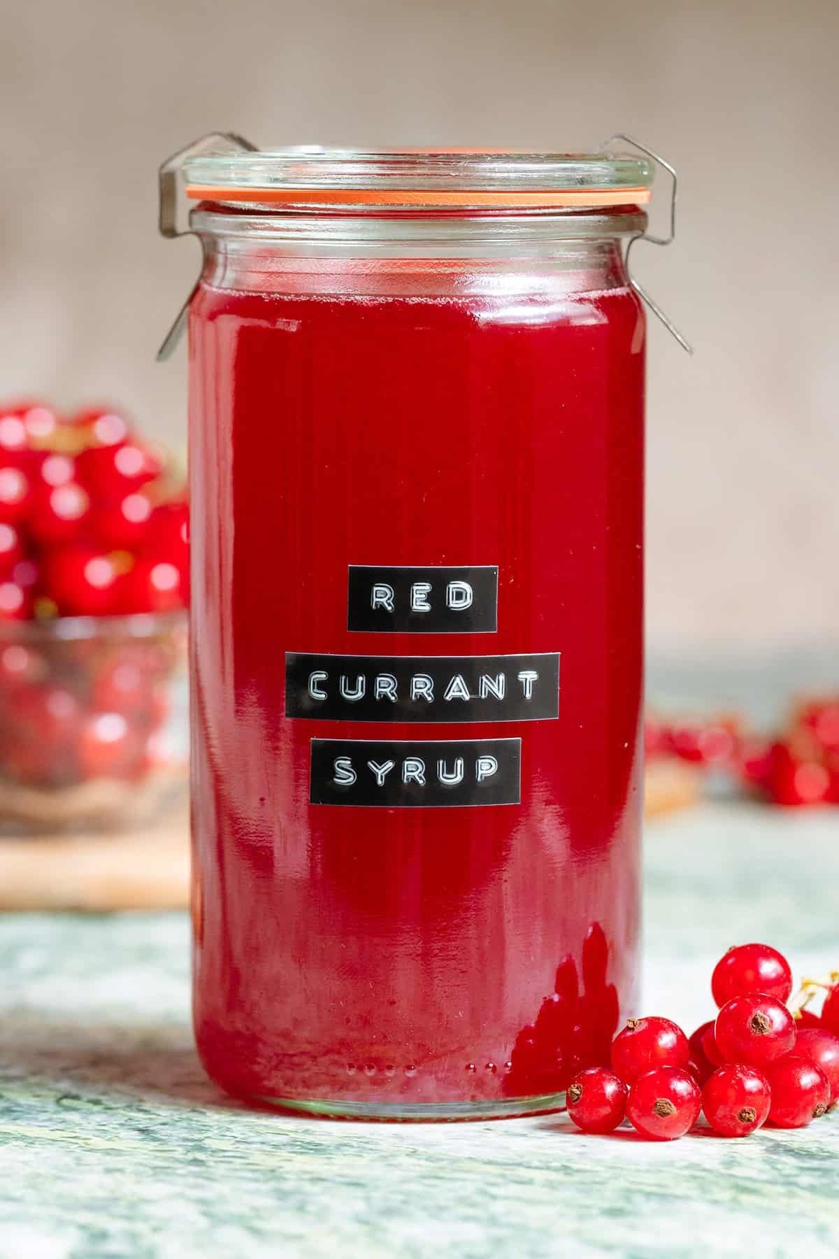 Vibrant red currant syrup in a tall glass jar with an embossed label on green marble backdrop with more red currants in the background.