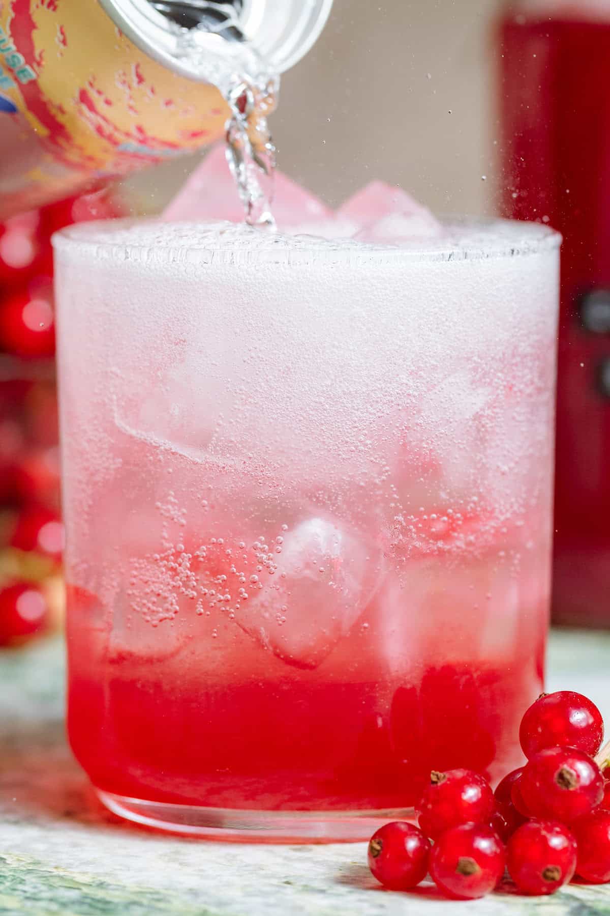 Sparkling water being poured into a short glass with ice and red currant syrup.