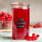 Vibrant red currant syrup in a tall glass jar with an embossed label on green marble backdrop with more red currants in the background.