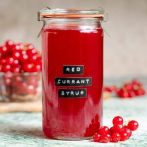 Vibrant red currant syrup in a tall glass jar with an embossed label on green marble backdrop with more red currants in the background.