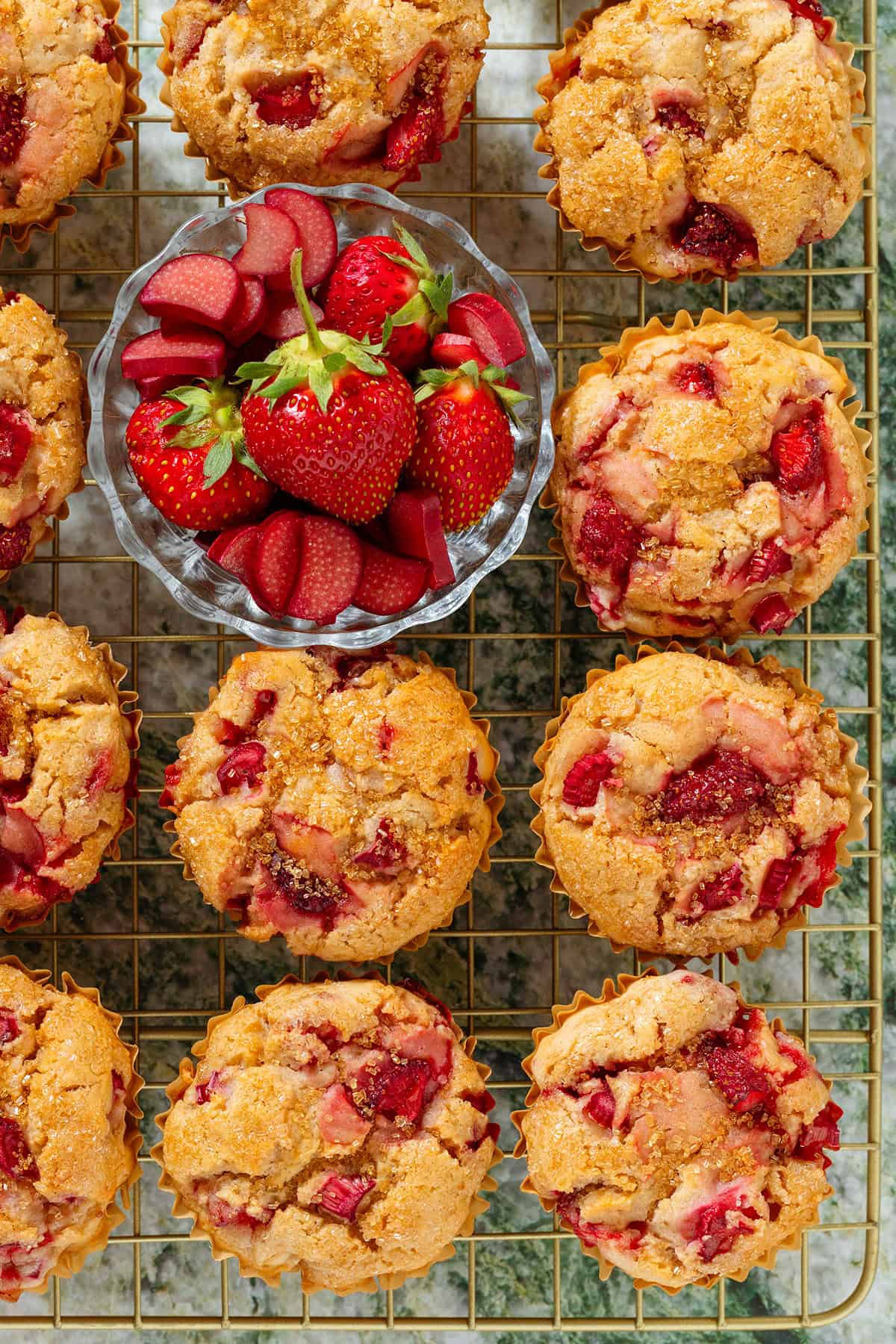 Strawberry rhubarb muffins on a gold wire rack with a small glass bowl of sliced rhubarb and fresh strawberries towards the top left of the frame.