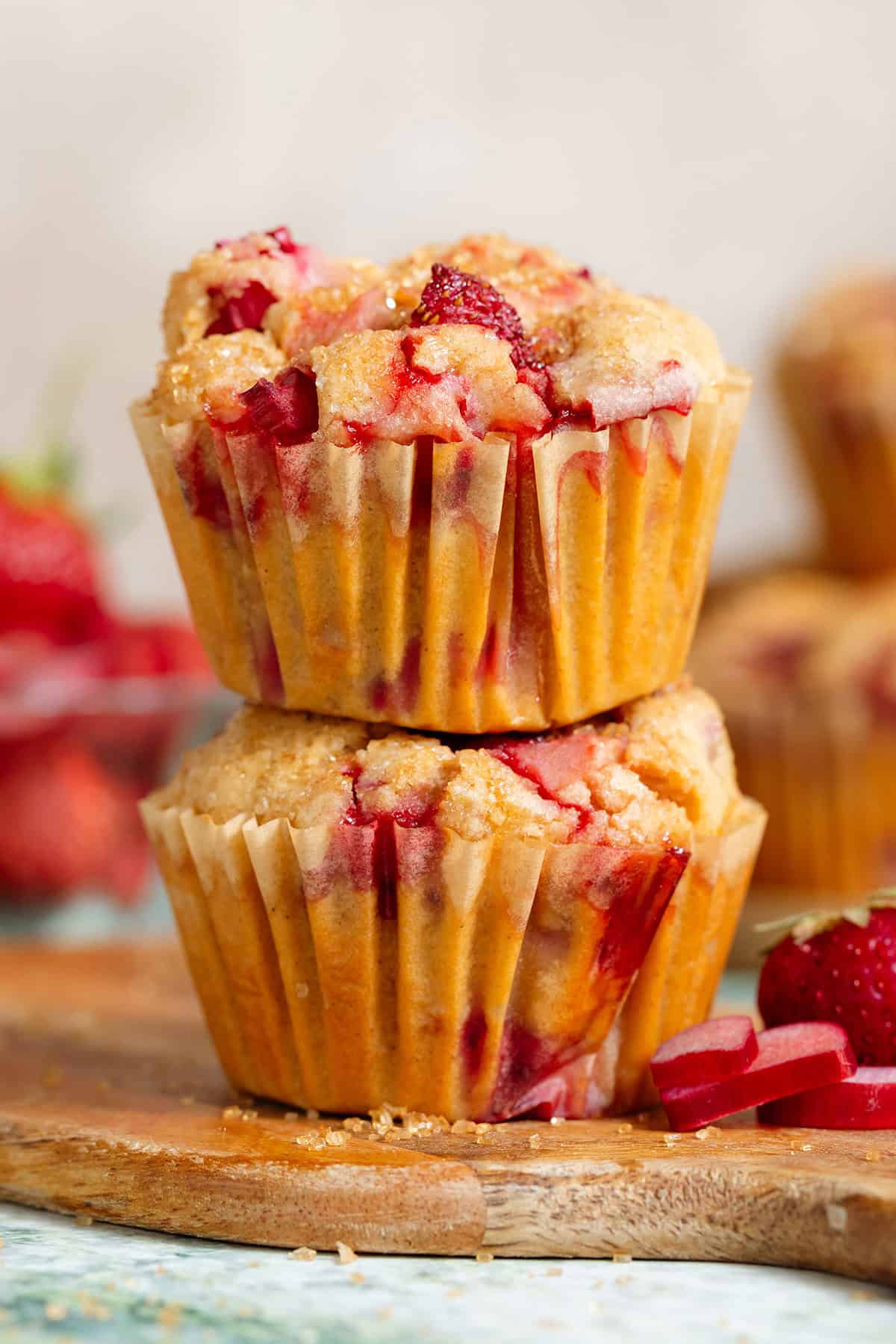 Two strawberry rhubarb muffins stacked on a small wooden cutting board with more sliced rhubarb, fresh strawberries, and muffins behind them.