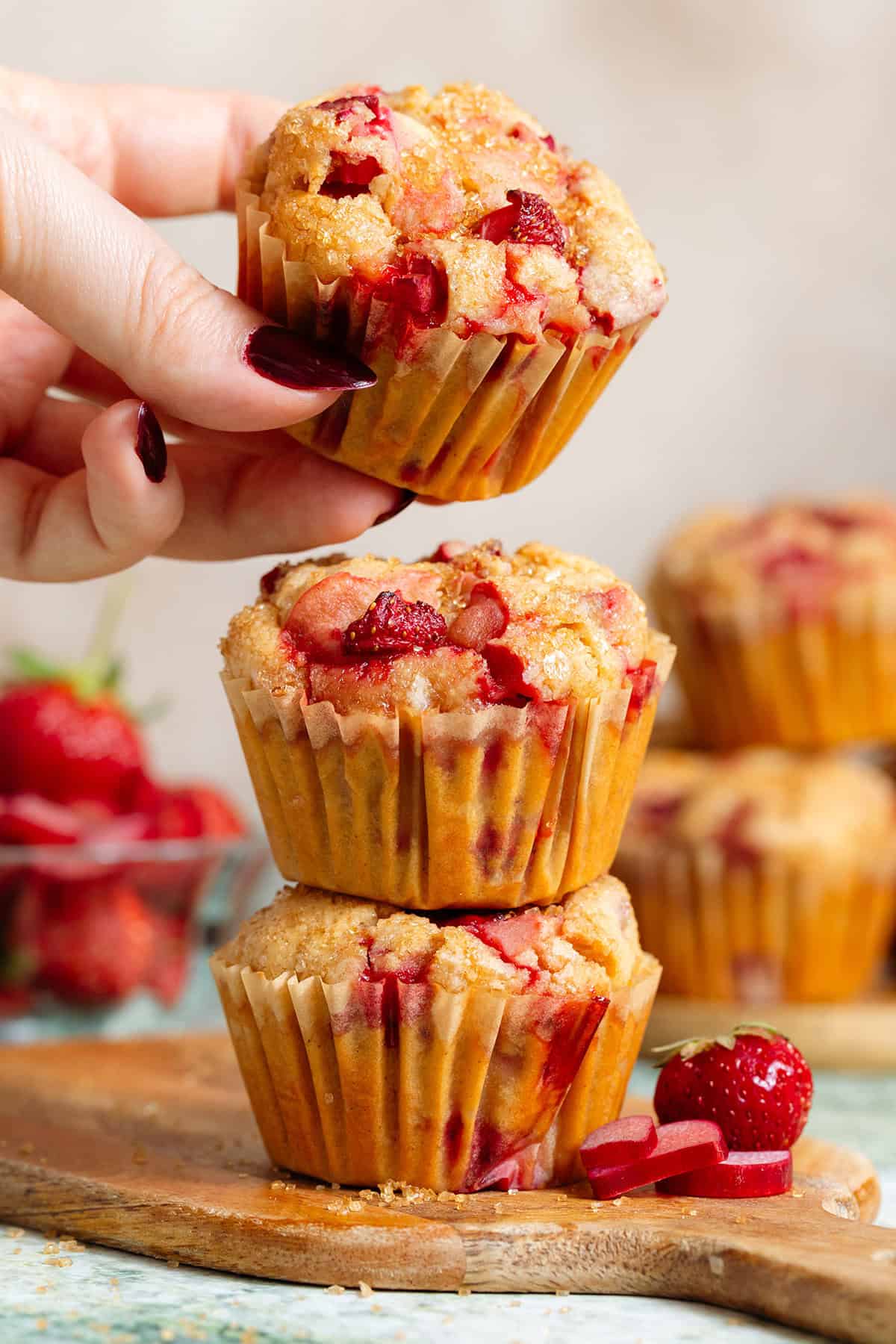 Three strawberry rhubarb muffins stacked on a small wooden cutting board with a hand grabbing the one on top.