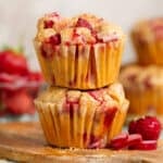 Two strawberry rhubarb muffins stacked on a small wooden cutting board with more sliced rhubarb, fresh strawberries, and muffins behind them.