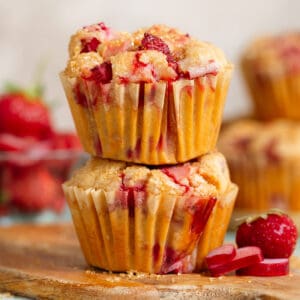 Two strawberry rhubarb muffins stacked on a small wooden cutting board with more sliced rhubarb, fresh strawberries, and muffins behind them.