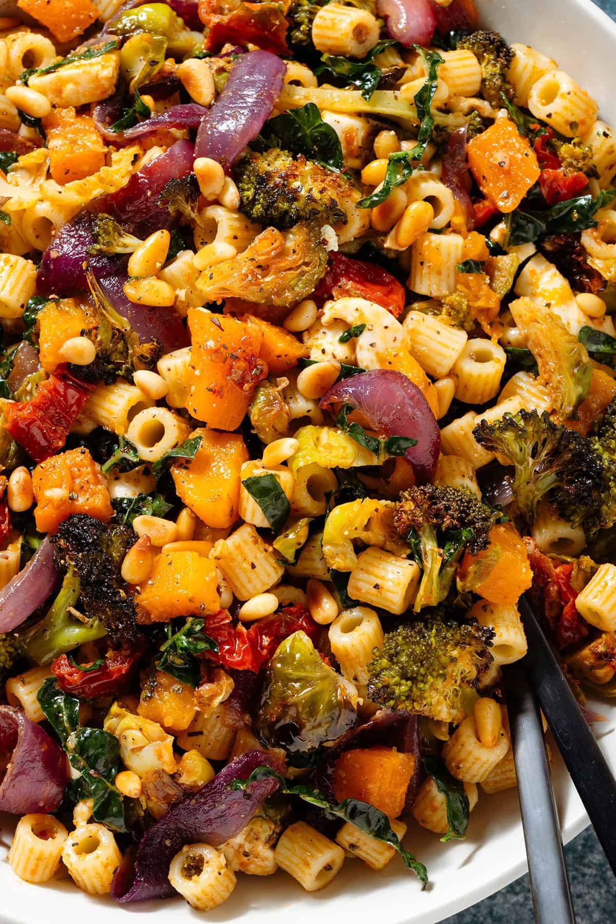 Fall pasta salad with mixed fall and winter vegetables in a large shallow bowl with black serving spoons inserted on the right.