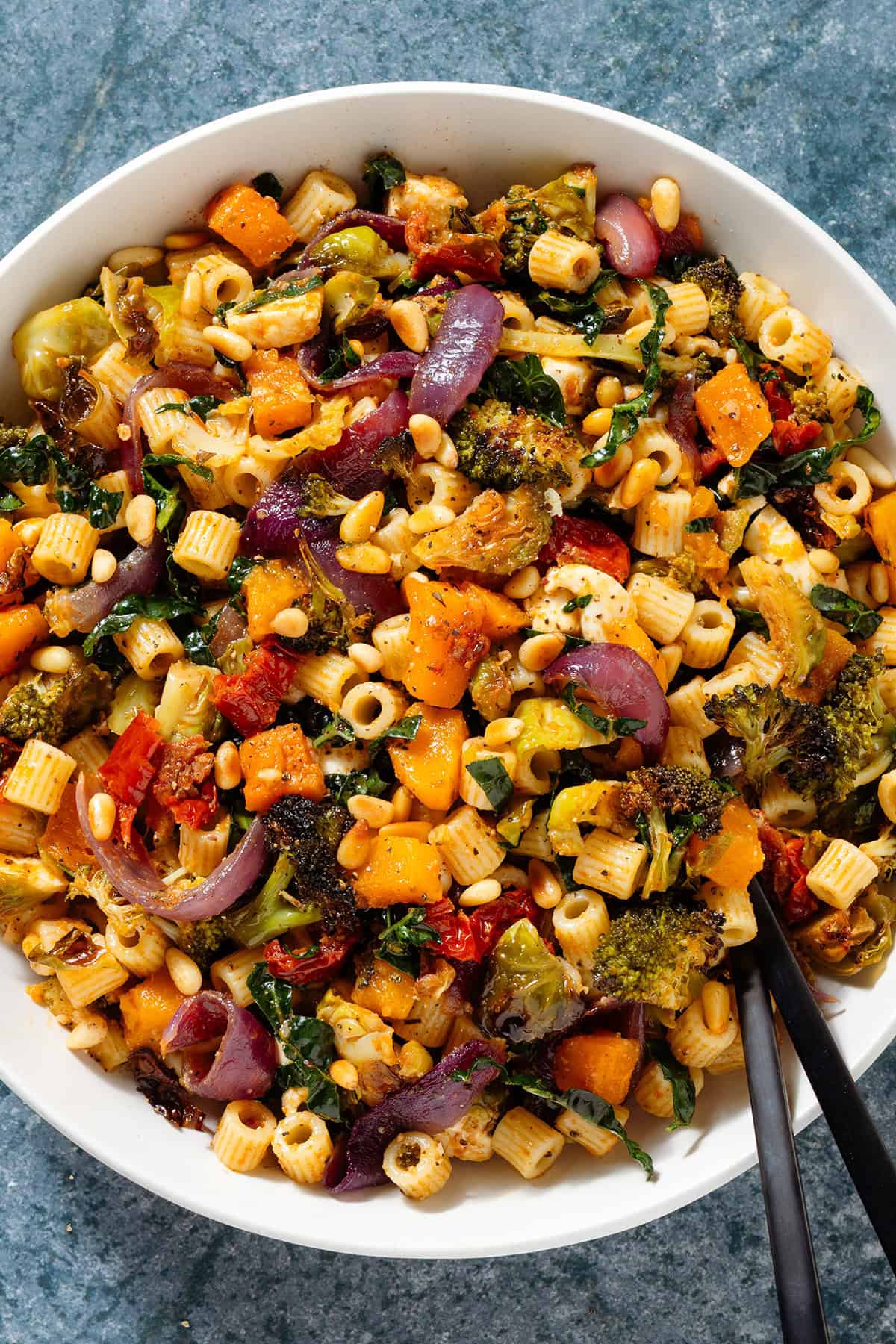 Fall pasta salad with mixed fall and winter vegetables, fresh mozzarella, pine nuts, and sundried tomato dressing in a large shallow bowl with black serving spoon.