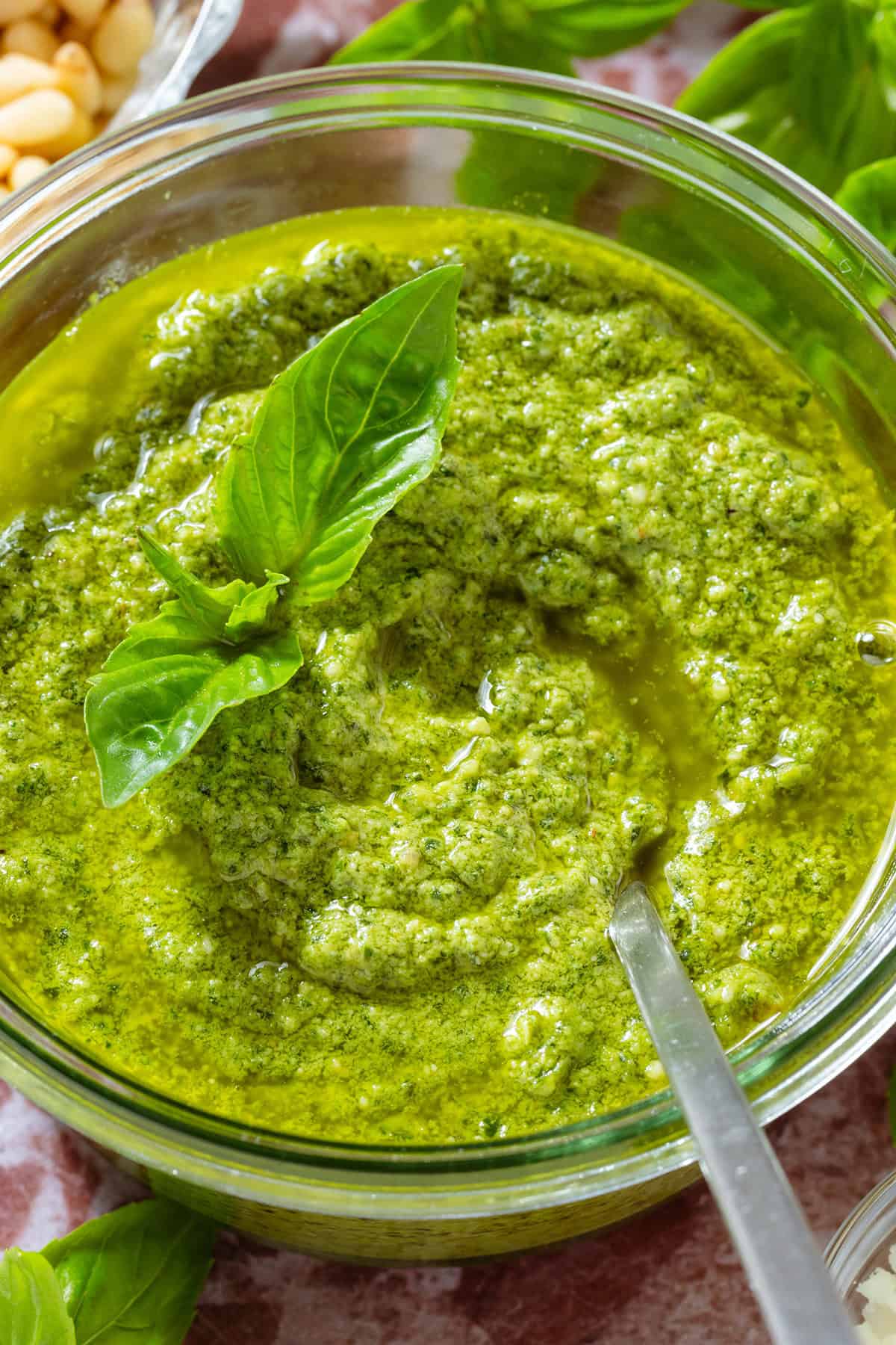 A glass jar with pesto garnished with fresh basil leaves and a spoon in the jar, surrounded with more basil, pine nuts, and pecorino.