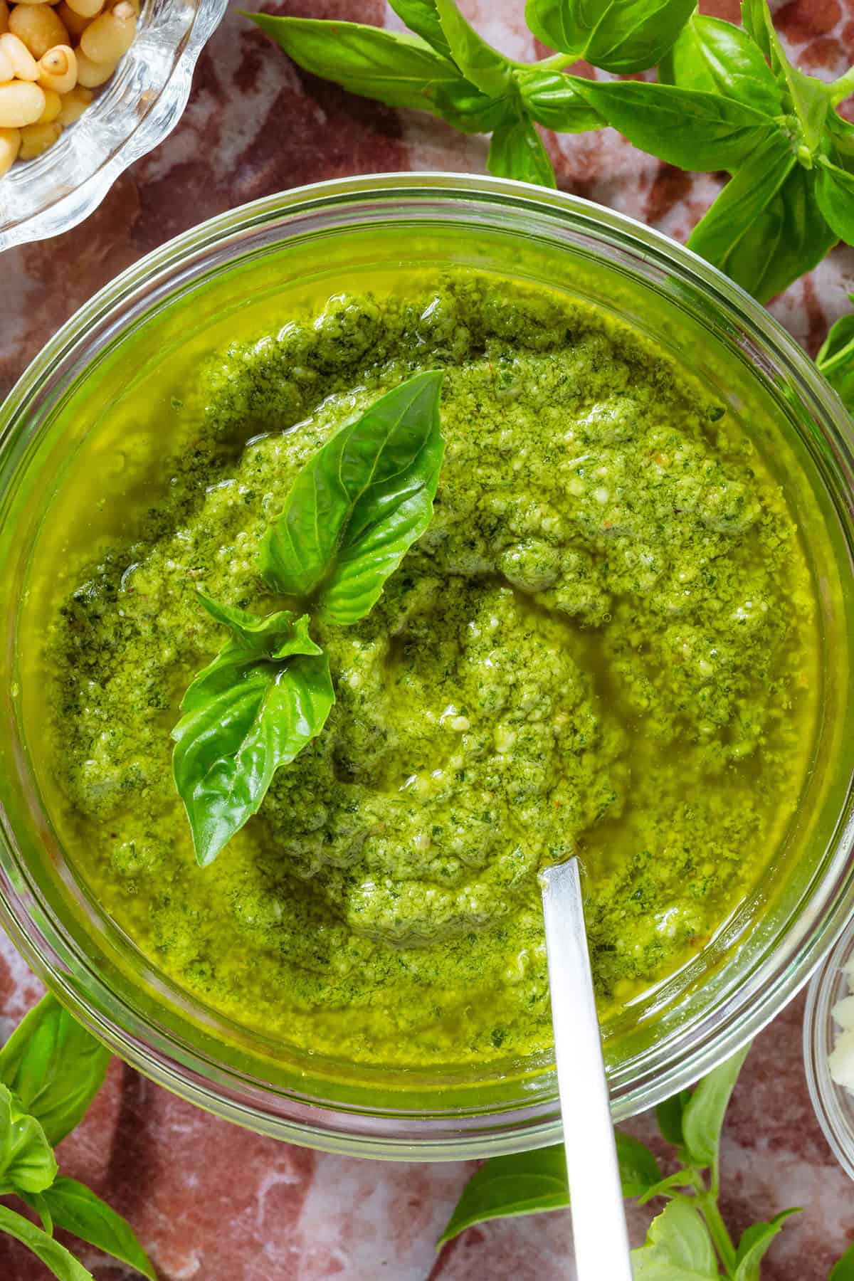 A glass jar with pesto garnished with fresh basil leaves and a spoon in the jar, surrounded with more basil, pine nuts, and pecorino.