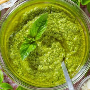 A glass jar with pesto garnished with fresh basil leaves and a spoon in the jar, surrounded with more basil, pine nuts, and pecorino.