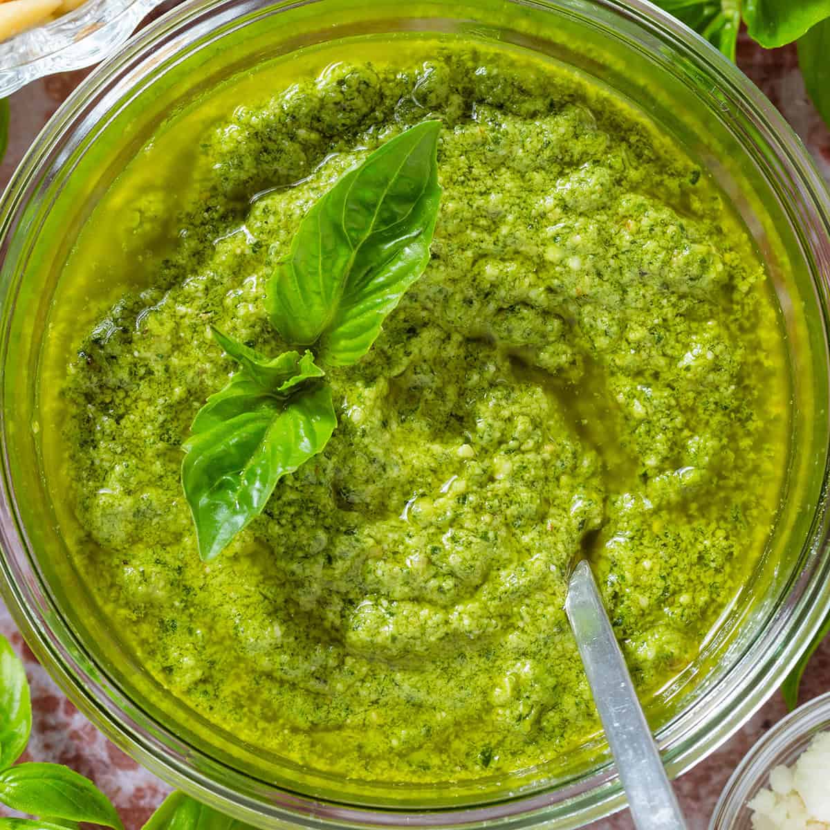 A glass jar with pesto garnished with fresh basil leaves and a spoon in the jar, surrounded with more basil, pine nuts, and pecorino.